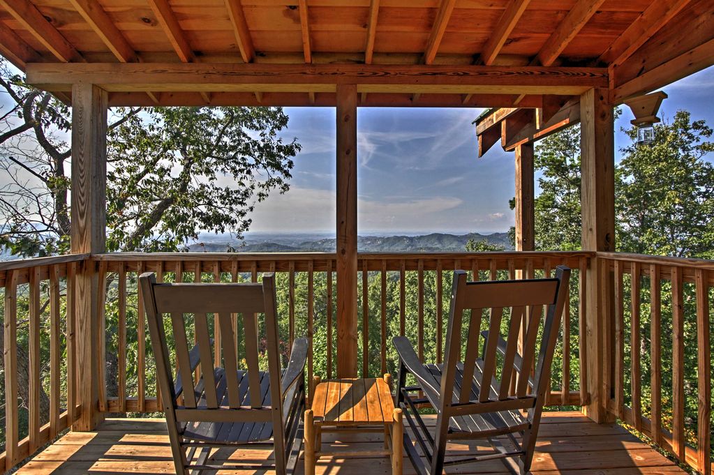 A balcony provides a serene outdoor space with wooden rocking chairs facing a wide, panoramic view of the surrounding forest and distant mountains. Natural light filters through the wooden beams overhead, creating a warm atmosphere while greenery flanks both sides.