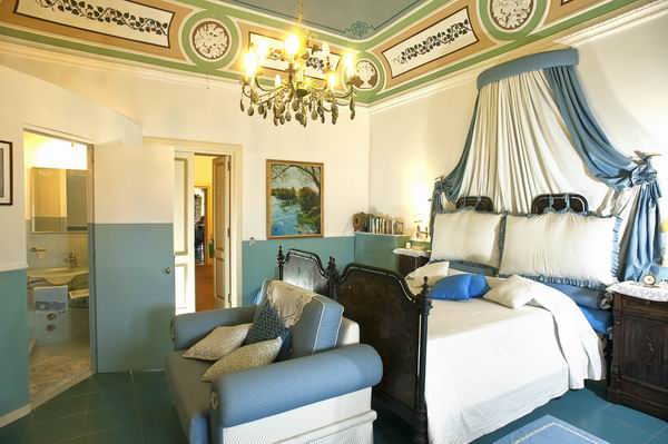A spacious room features a king-size bed adorned with decorative bedding and a large headboard. A comfortable seating area includes a light blue sofa with patterned cushions. The ceiling boasts intricate designs, and a modern ensuite bathroom is visible through a partially open door.