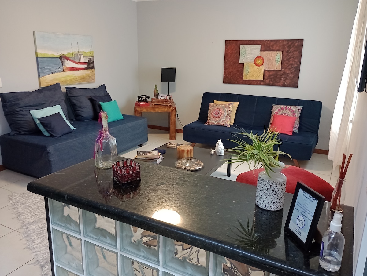 A living area is featured with two sofas, decorative pillows, and a coffee table. A painting of a boat decorates the wall above a wooden table. A glass block wall separates the kitchen space, adding to the open ambiance.