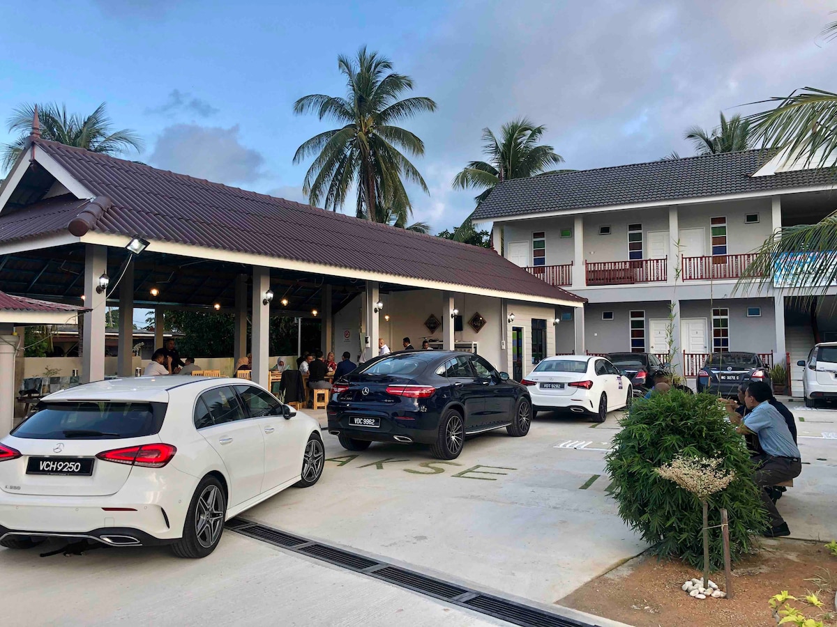 The exterior of the homestay features a spacious parking area accommodating multiple cars, surrounded by lush greenery. A two-story building is visible, with a sloped roof and a welcoming porch area, providing access to individual guest rooms.