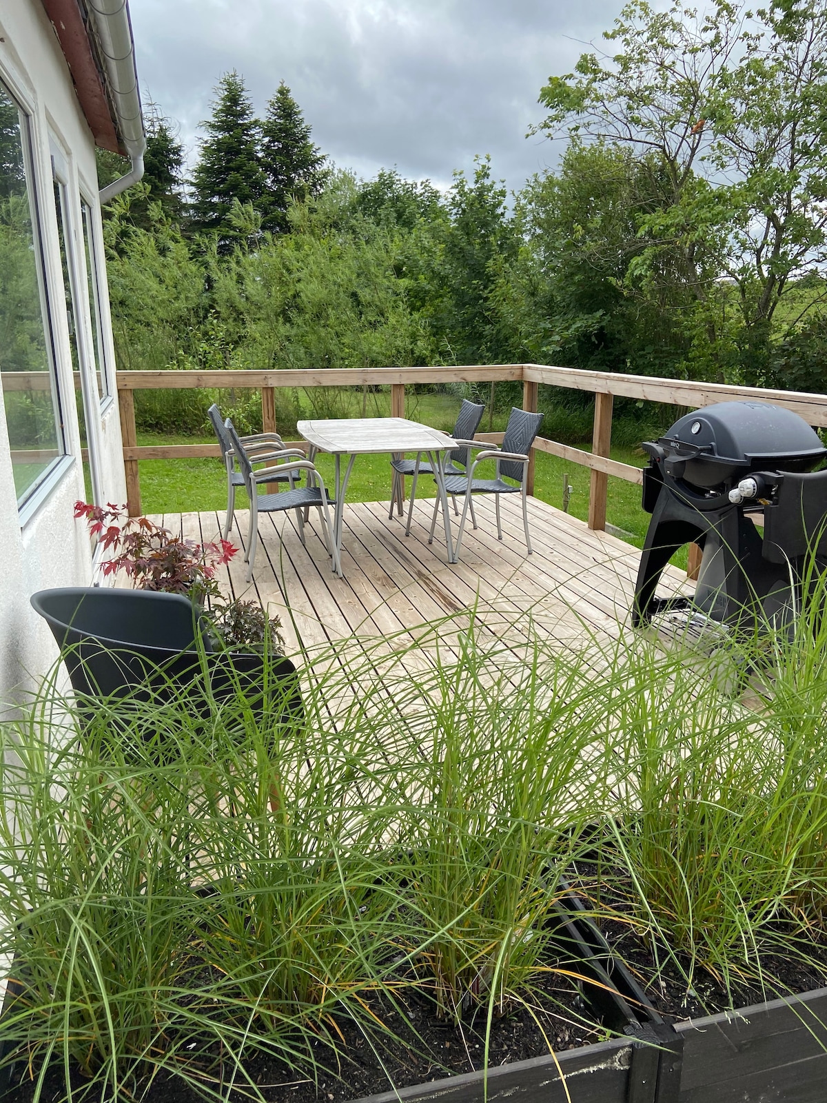 A wooden terrace area is shown, featuring a round table surrounded by four chairs, alongside a gas grill. Green grass and shrubs frame the space, with trees in the background, creating a serene outdoor setting for relaxation.