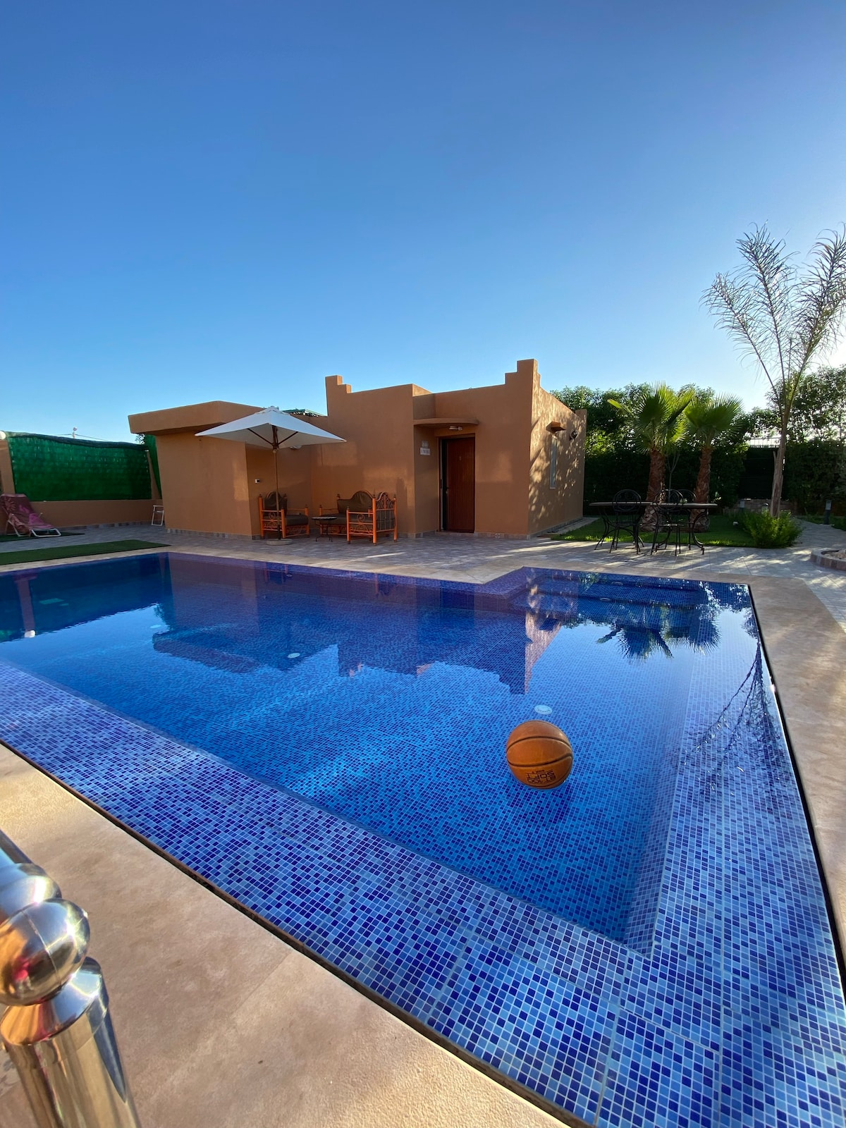 The outdoor area features a glistening pool surrounded by a tiled patio, with lounge chairs beneath an umbrella. Lush greenery and palm trees border the space, providing a serene environment. A basketball floats gently on the water's surface, hinting at recreational options.