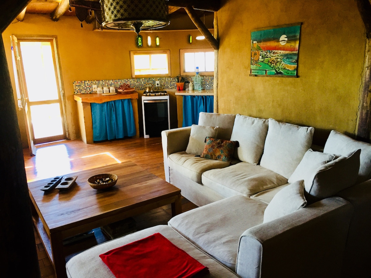 A spacious living area features a comfortable L-shaped sofa in a neutral tone, complemented by a wooden coffee table. A vibrant piece of artwork hangs on the wall, and the kitchen area showcases colorful cabinetry and appliances, illuminated by natural light from large windows.
