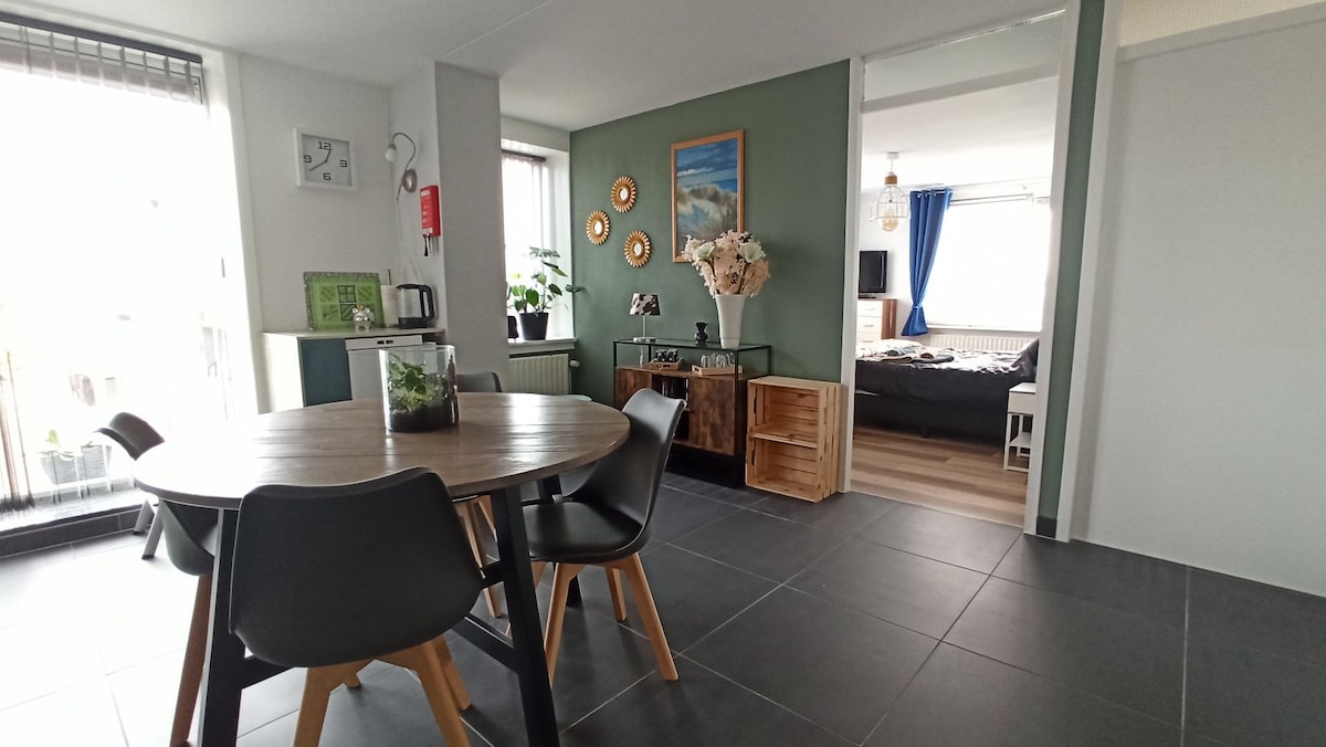 A modern dining area is featured with a round table and black chairs. The adjacent living space showcases a green accent wall and decorative elements. A well-lit bedroom is visible in the background, with large windows allowing natural light into the room.