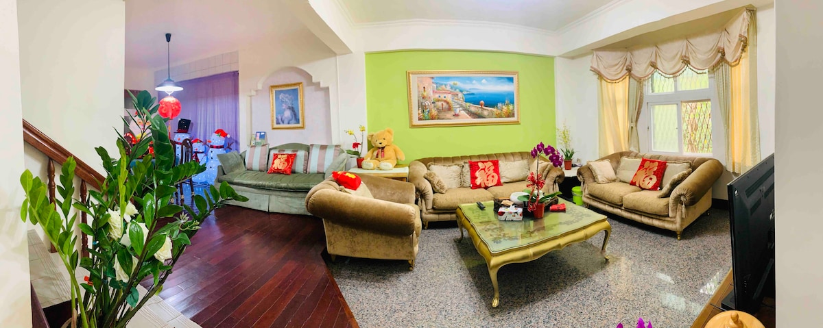 The spacious living area features comfortable seating arrangements with plush sofas adorned with colorful cushions. A glass coffee table is centered among decorative plants and flowers. Natural light is filtered through large windows draped with soft curtains, enhancing the inviting atmosphere.