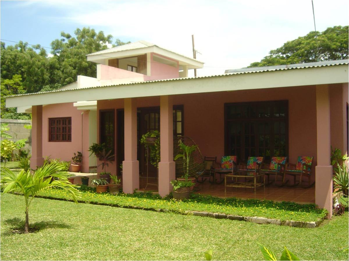 A charming house is set amidst a lush green garden, featuring a covered porch with colorful seating. Large windows allow natural light into the interior, while potted plants enhance the inviting atmosphere. The roofline provides shade, complementing the overall serene setting.