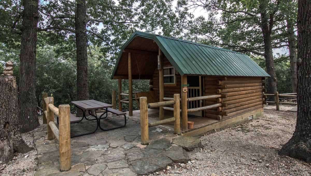 A rustic cabin with a green metal roof is surrounded by trees, featuring a front porch with a picnic table. The cabin's wooden exterior complements the natural stone flooring. Wooden railings are visible on the porch, enhancing the inviting outdoor space.
