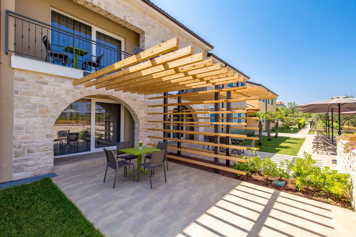 An outdoor seating area features a wooden pergola, providing shade over a green table with four chairs. Lush landscaping surrounds the area, enhancing the tranquil setting. The façade of the house showcases stone details and large windows, inviting natural light into the space.
