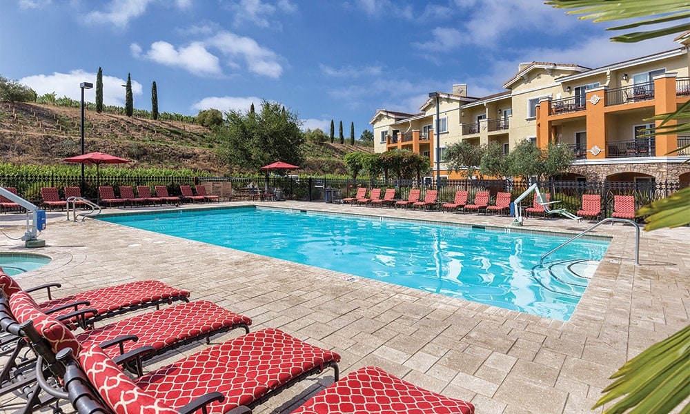 A spacious outdoor pool area is surrounded by ample lounge chairs with red cushions. The pool is framed by stone pavers, and a scenic hillside with vineyards is visible in the background. Umbrellas provide shade, enhancing the relaxing atmosphere.