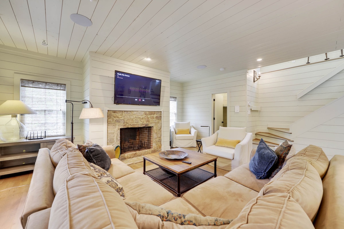 A welcoming living area features a large, curved sectional sofa arranged around a wooden coffee table. A flat-screen TV is mounted above a stone fireplace, with two white armchairs providing additional seating. Natural light filters through the large windows, enhancing the open and airy ambiance.