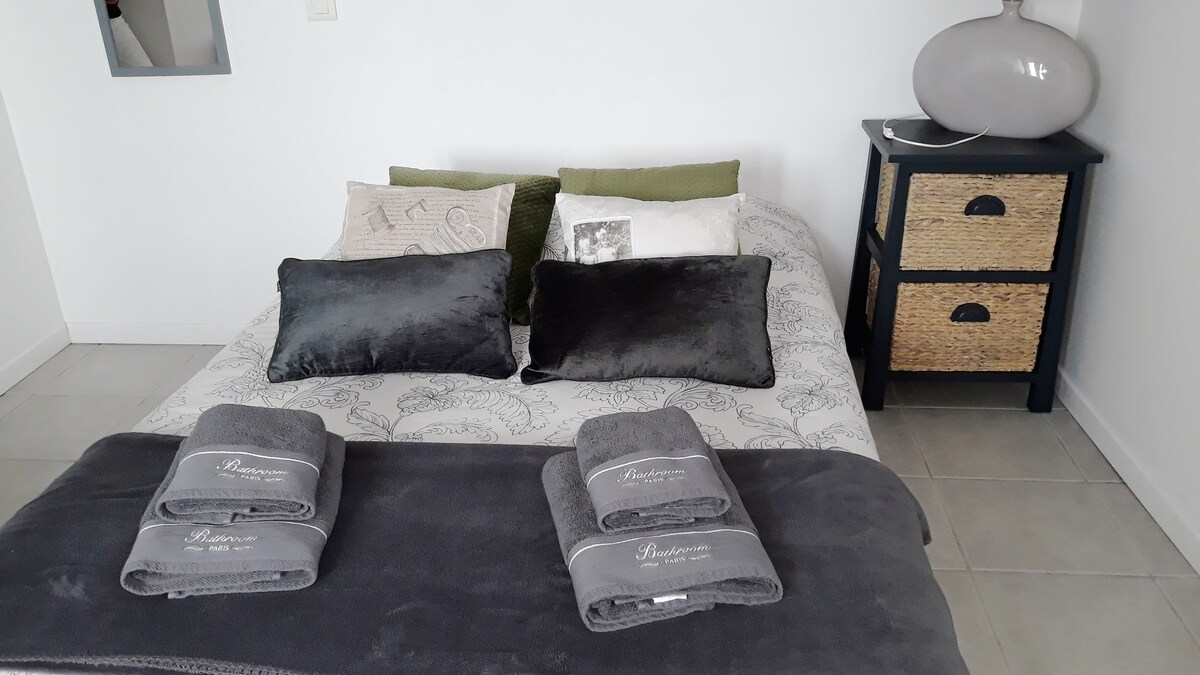 A comfortable bed is presented with plush pillows and neatly arranged towels. The bedding features a subtle design, complemented by green throw pillows. A bedside table with a textured surface and a decorative lamp is visible, alongside a woven storage basket.