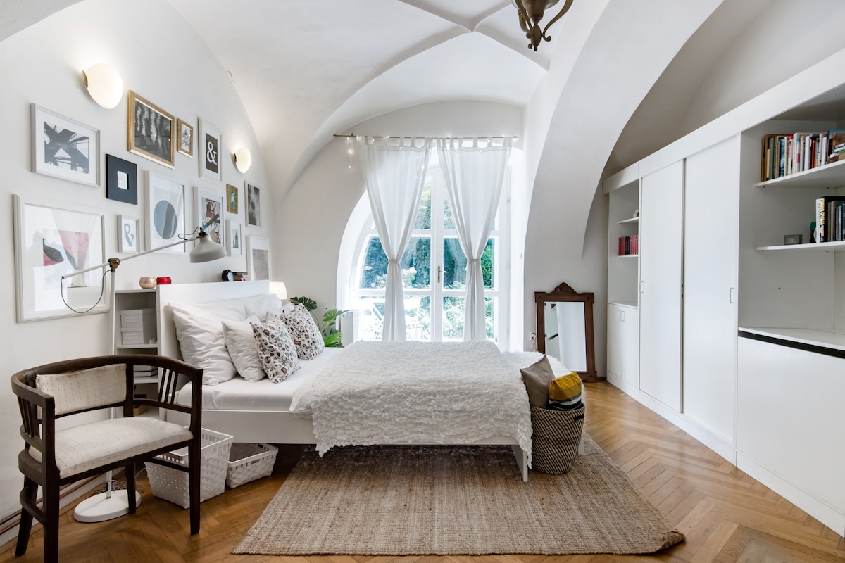 A spacious studio bedroom features a white bed dressed with a textured blanket, complemented by patterned pillows. A set of double glass doors leads to the garden, allowing natural light to fill the room. A wooden chair and woven basket add functional decor.