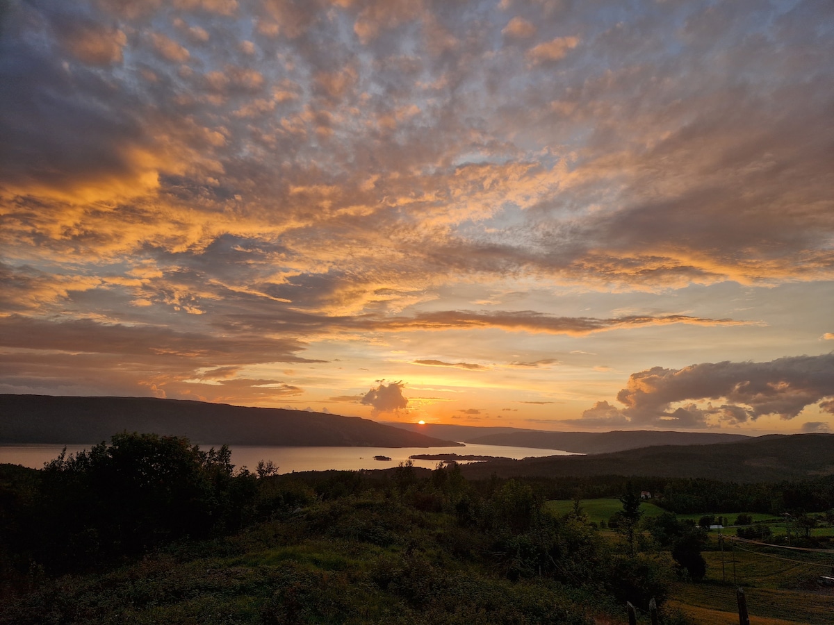 A panoramic view showcases a sunset over Randsfjorden, with hues of orange and pink illuminating the clouds. The surrounding landscape includes lush greenery and fields, creating a serene backdrop for the tranquil scene.