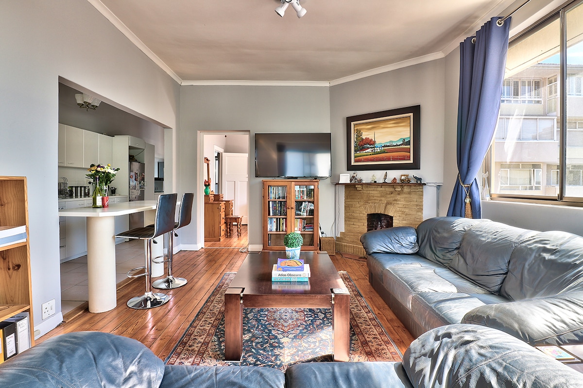 A spacious living room is presented with original wood floors and high ceilings. A large sectional sofa surrounds a coffee table atop a decorative rug. A fireplace is visible, and natural light streams through large windows adorned with blue curtains, illuminating the modern decor.