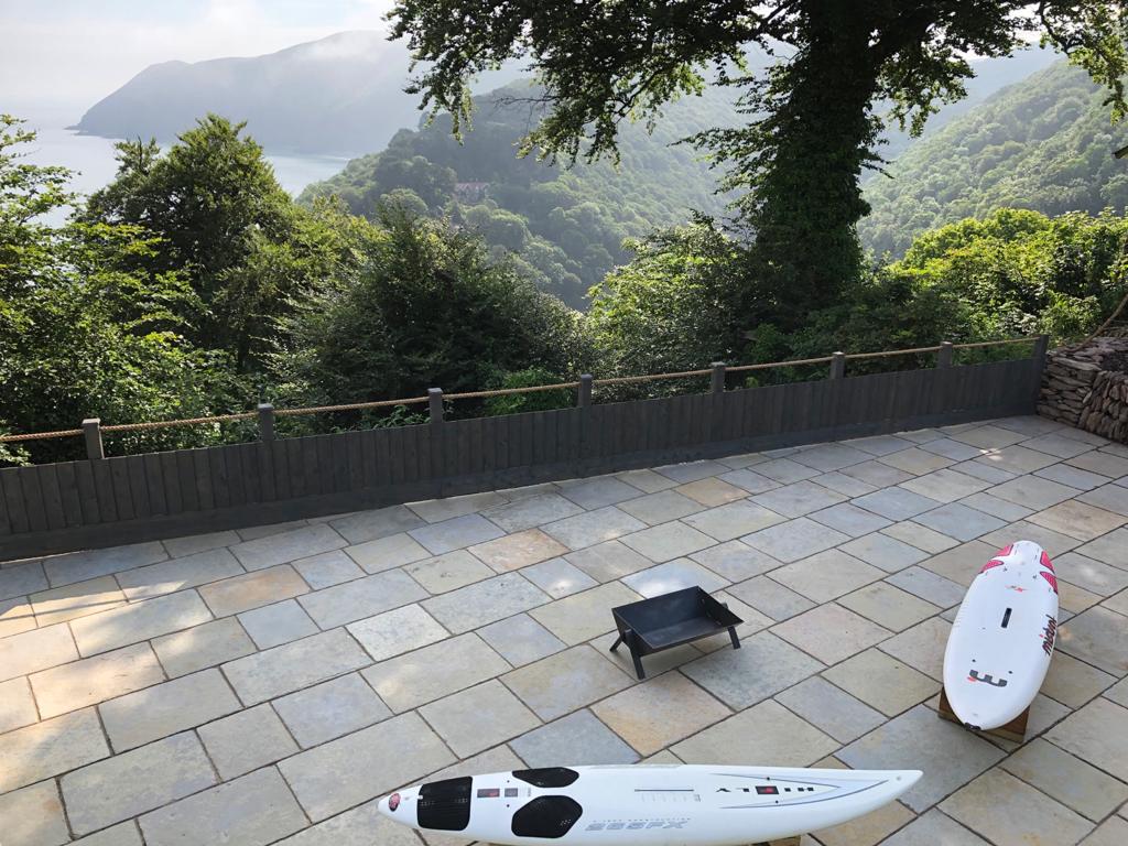 A spacious limestone patio is presented, featuring two kayaks positioned beside a fire pit. Lush greenery surrounds the area, and a scenic view of the coastline is visible in the distance, creating a serene outdoor environment.