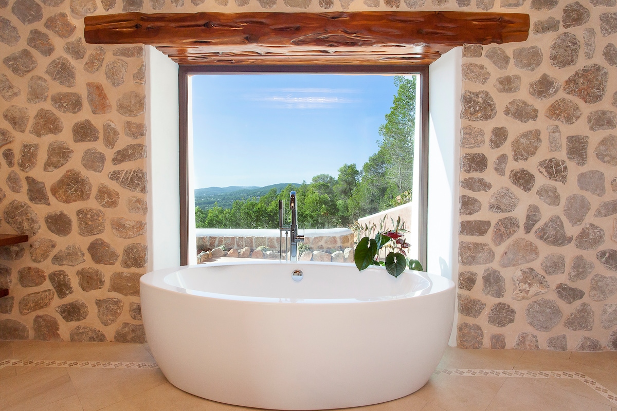 A freestanding soaking tub is positioned by a large window, providing a view of the lush greenery outside. Natural stone walls frame the space, while a wooden beam enhances the rustic charm. A potted plant adds a touch of greenery to the serene setting.
