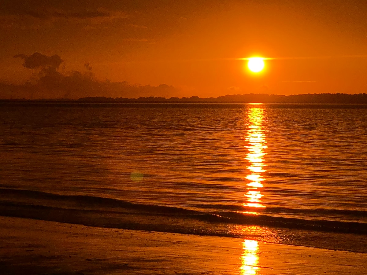 A serene beachfront scene is captured during sunset, with the sun reflecting off the calm water. The horizon features a warm orange glow, merging with soft silhouettes of distant land. Gentle waves lightly lap against the sandy shore, creating a peaceful atmosphere.
