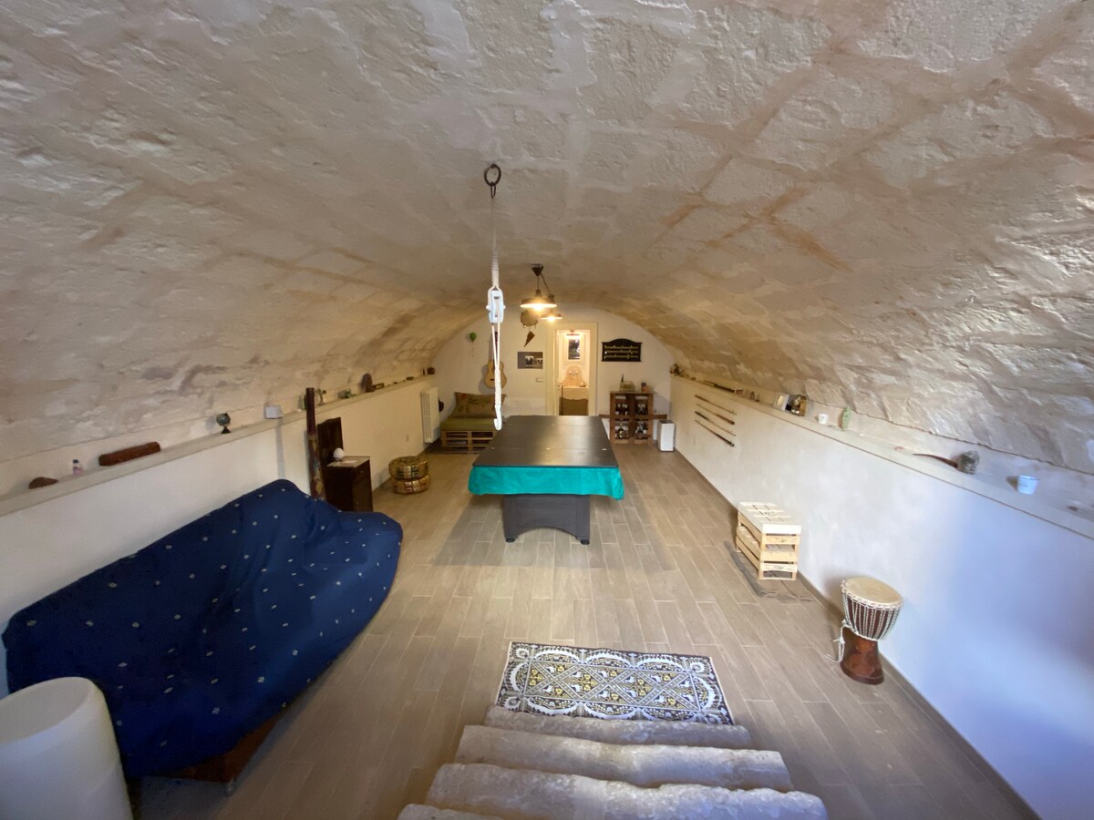 An underground space features an arched stone ceiling with warm lighting. A green billiard table is centrally placed, surrounded by simple furnishings including a blue couch and decorative items. Steps lead up to an entrance visible in the background, enhancing the inviting atmosphere.