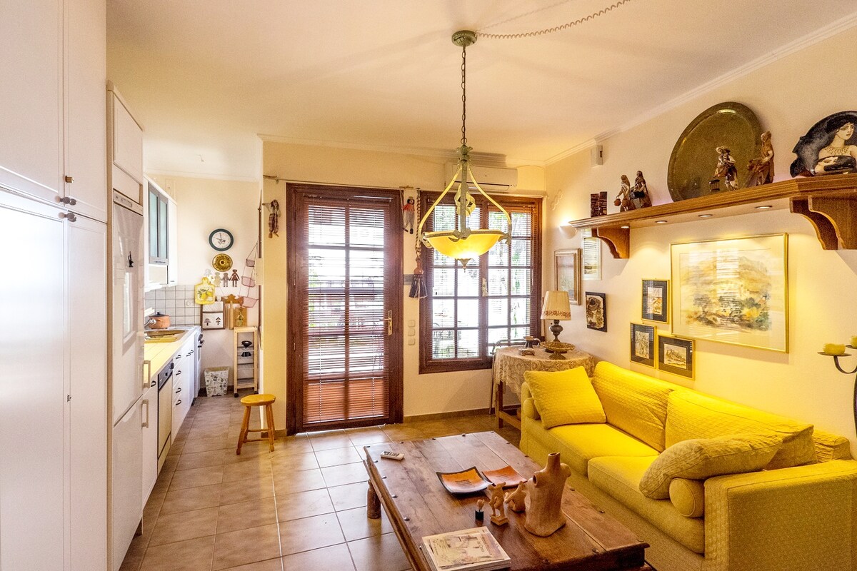 A cozy living area features bright yellow furnishings and warm wooden accents. A wooden coffee table is centered in front of a comfortable sofa, creating a welcoming space. Natural light pours in through large windows, illuminating the open kitchen and decorative wall art throughout the room.