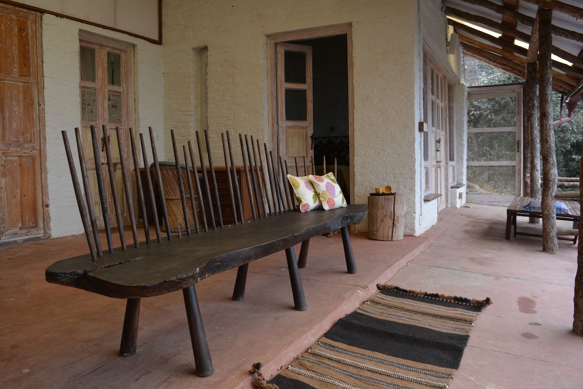 A rustic seating area features a long wooden bench with distinctive slat backrests and a decorative pillow. The space is complemented by a woven rug and showcases natural materials, allowing for a connection with the surrounding forest.