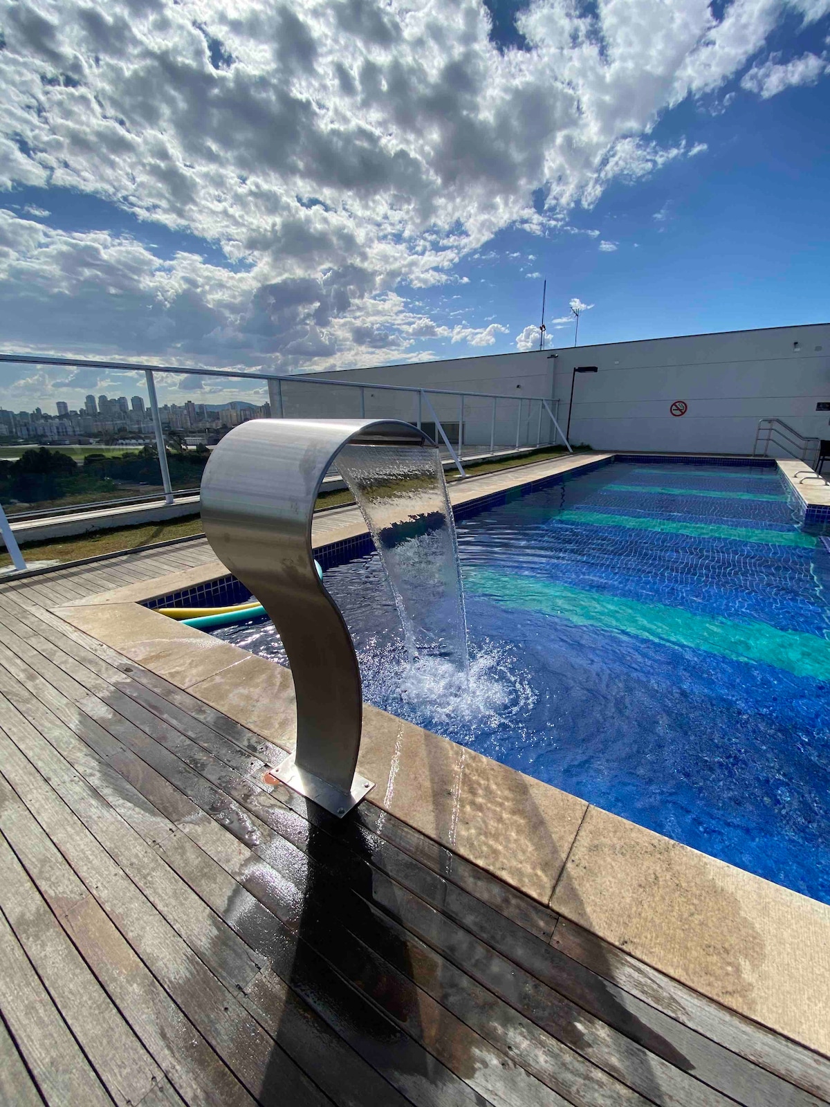 A rooftop pool is showcased, featuring a sleek waterfall feature that cascades into the clear blue water. The surrounding deck is made of wooden planks, offering a warm contrast to the pool's vibrant colors. A city skyline can be seen in the background under a partly cloudy sky.