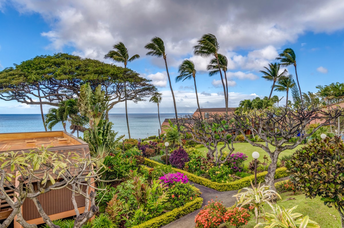 Lush gardens filled with vibrant flowers are visible, framed by palm trees swaying in the breeze. The view extends to the ocean, showcasing a serene backdrop against a partly cloudy sky. Pathways meander through the greenery, inviting exploration of the beautiful grounds.