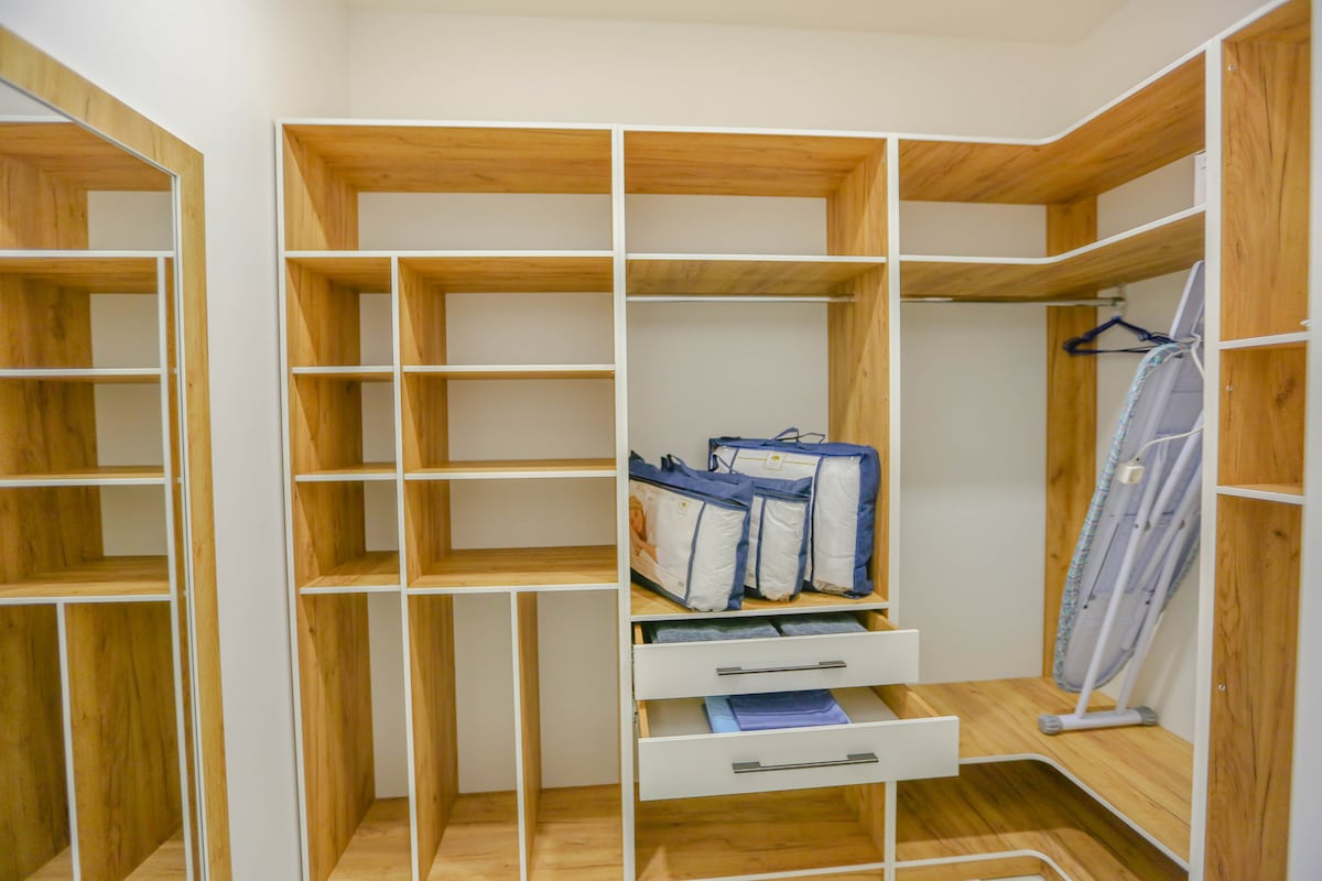 A spacious closet area features multiple wooden shelves and drawers, providing ample storage. An ironing board is positioned neatly against one side, while two storage bags rest on a shelf below. Full-length mirrors line one wall, enhancing the room's brightness and openness.