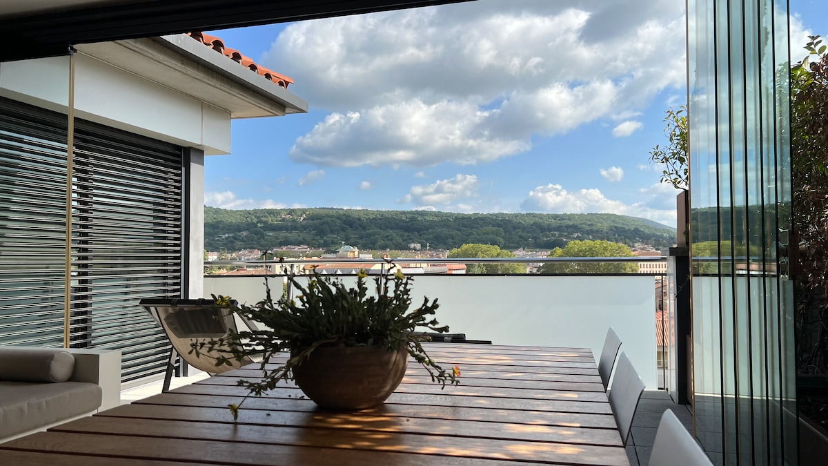 A spacious terrace is displayed, featuring a wooden dining table surrounded by comfortable seating. Decorative plants sit on the table, and expansive views of the landscape and clouds are visible in the background, creating an inviting space for relaxation.