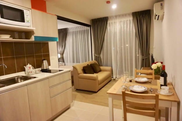 An open-plan living area is visible, featuring a well-equipped kitchen with modern appliances and a cozy seating area. A small dining table is set for two, adorned with dishes and wine. Large windows allow natural light to flow in, with curtains neatly draped.