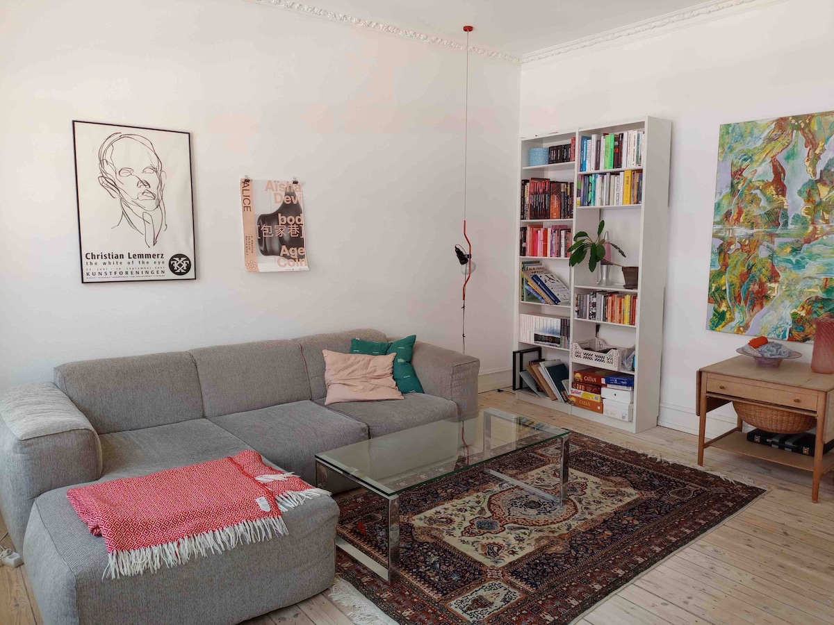 A spacious living area features a light grey sectional sofa adorned with a colorful throw and decorative cushions. A glass coffee table is placed on a patterned rug, complemented by a white bookcase filled with books. Artwork adds a vibrant touch to the wall.