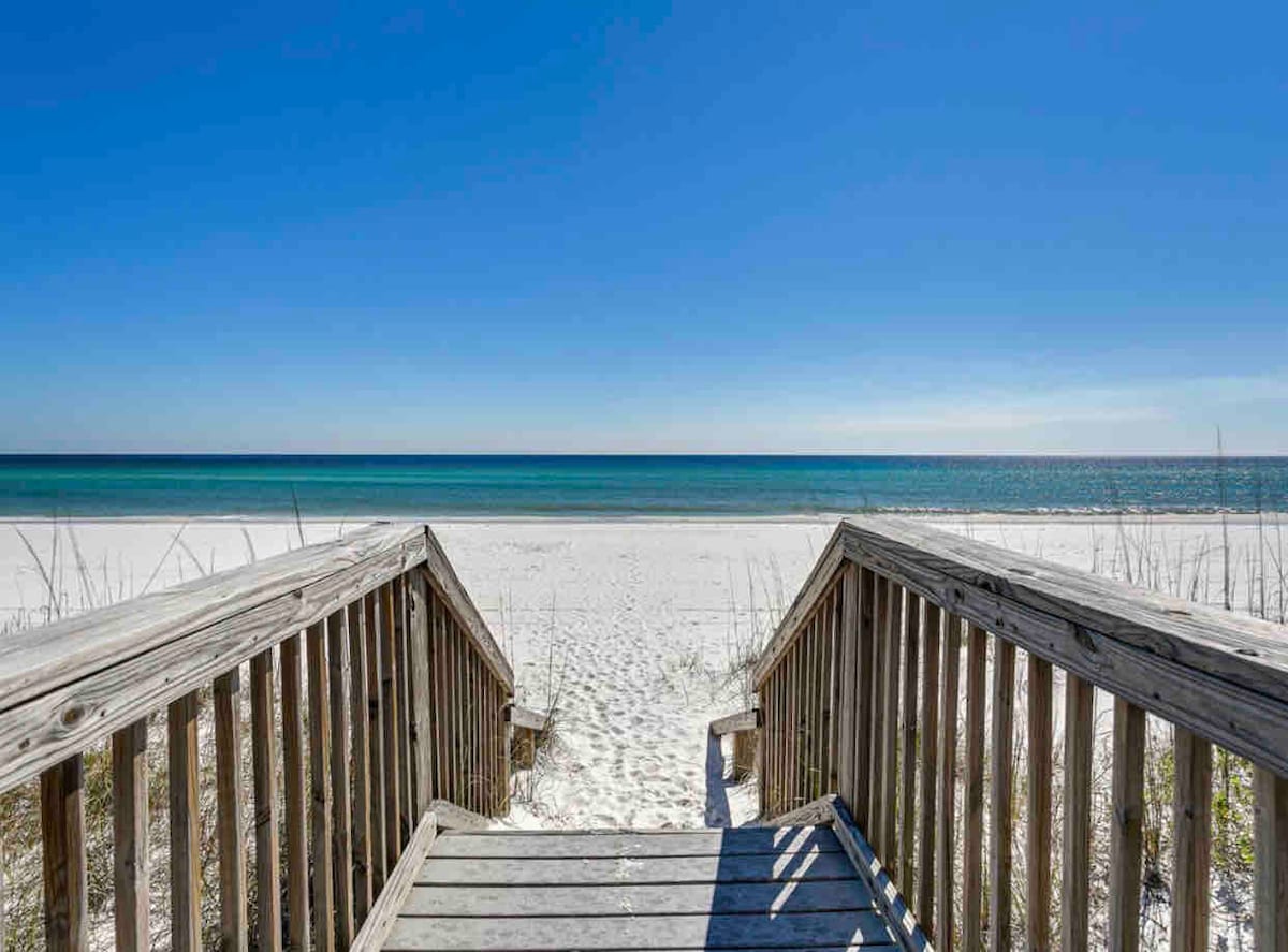 A wooden boardwalk leads directly to a tranquil beachfront. The soft, white sand stretches along the shoreline, meeting gentle blue waves under a clear, bright sky. Sparse grasses frame the sand, contributing to the natural coastal ambiance.