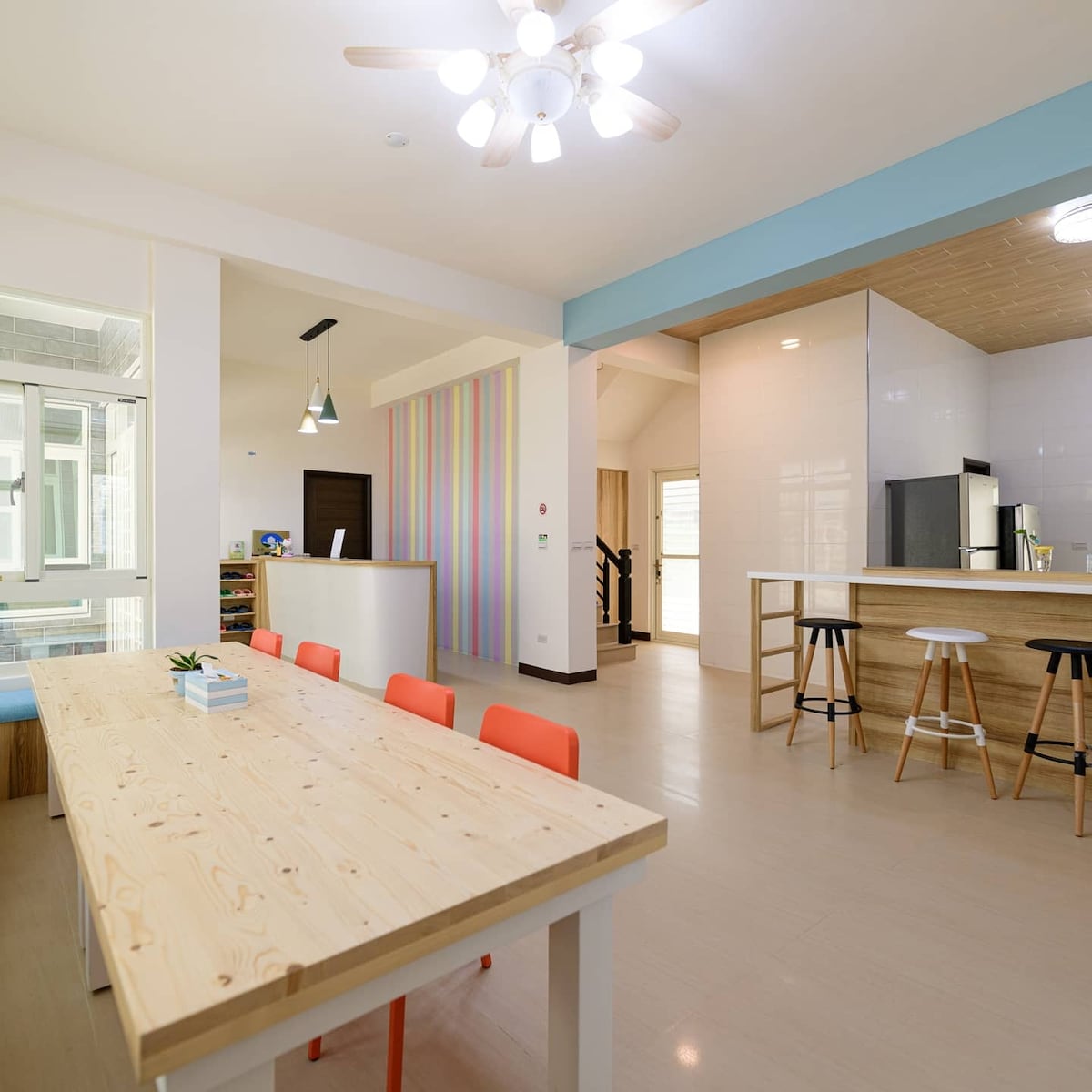 An open-plan living area features a large wooden dining table surrounded by colorful chairs. Bright walls and a light ceiling create an airy atmosphere. The kitchen area includes bar stools, and natural light enhances the overall spaciousness.