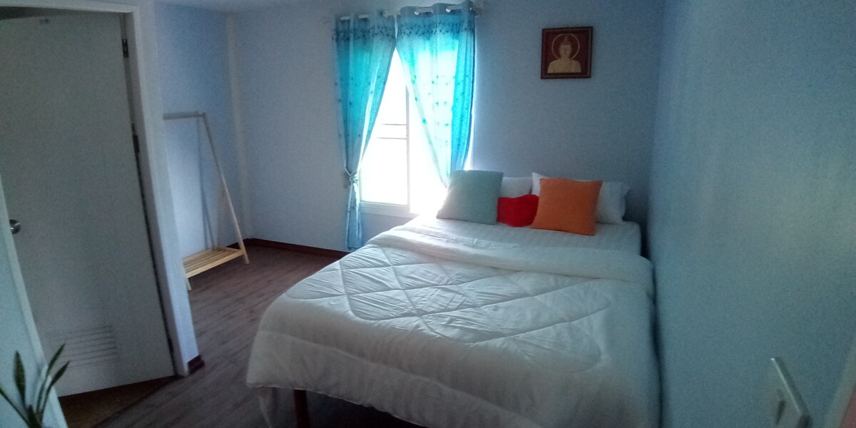 A bright sleeping area features a spacious bed layered with a white quilt and colorful accent pillows. Natural light floods in through a window adorned with sheer blue curtains. A wooden rack stands against the wall, adding functionality to the room's design.