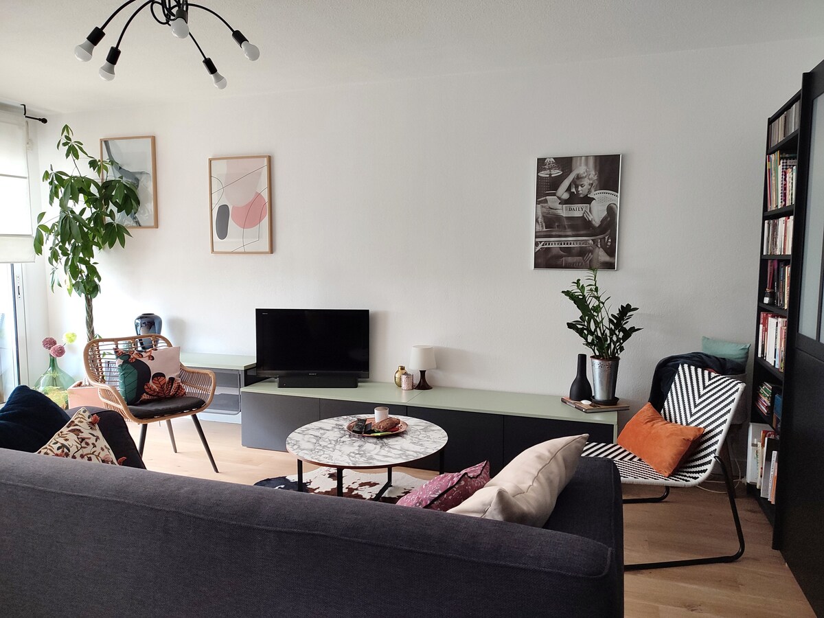 A bright living room features a comfortable gray sofa and a stylish armchair. A round marble coffee table centers the space, while a TV is mounted on a sleek cabinet. Decorative plants and wall art add a touch of character to the inviting environment.