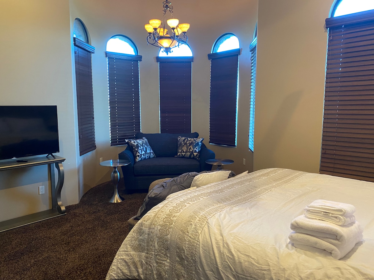A comfortable guest bedroom showcases a spacious bed with neatly arranged towels. A seating area features a sofa and two side tables, complemented by large windows with wooden blinds that allow natural light to filter in. A television is positioned for easy viewing.