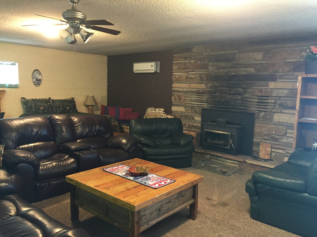 A cozy living space features a combination of dark leather sofas arranged around a rustic wooden coffee table. A stone fireplace serves as a focal point, and a subtle ceiling fan provides air circulation. Natural light enters through a small window, enhancing the inviting atmosphere.