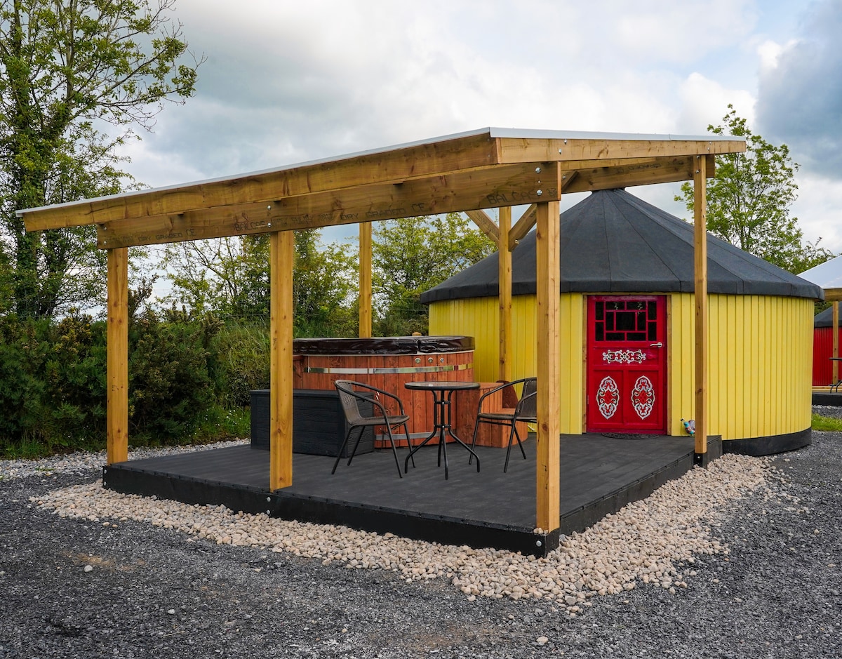 An outdoor deck features a covered area beside a vibrant yellow yurt. A wooden hot tub is visible, surrounded by stony landscaping. A black table and two chairs are arranged on the deck, providing a space for relaxation and enjoying the scenery.