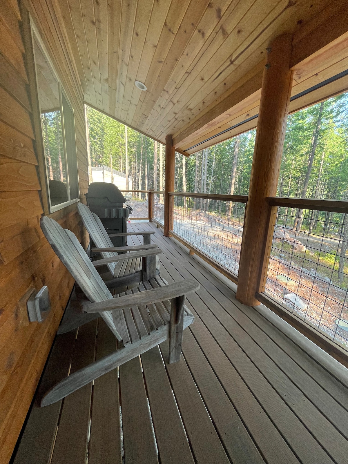 A covered deck is viewable, featuring two wooden chairs positioned beside a BBQ grill. Tall trees rise in the background, providing a serene forest atmosphere. The deck's wooden flooring complements the cabin's rustic charm, inviting relaxation amid nature.