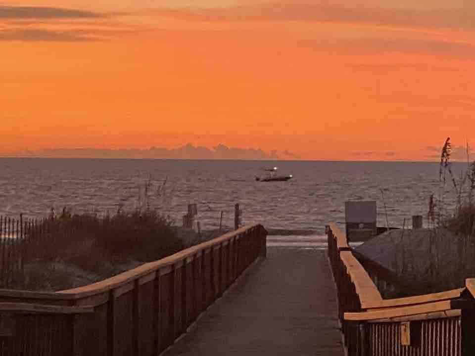 A boardwalk leads towards the ocean, framed by grassy dunes on either side. The sky displays a blend of warm orange and soft purple hues during sunset. A boat is seen cruising on the water, while gentle waves lap against the shore.