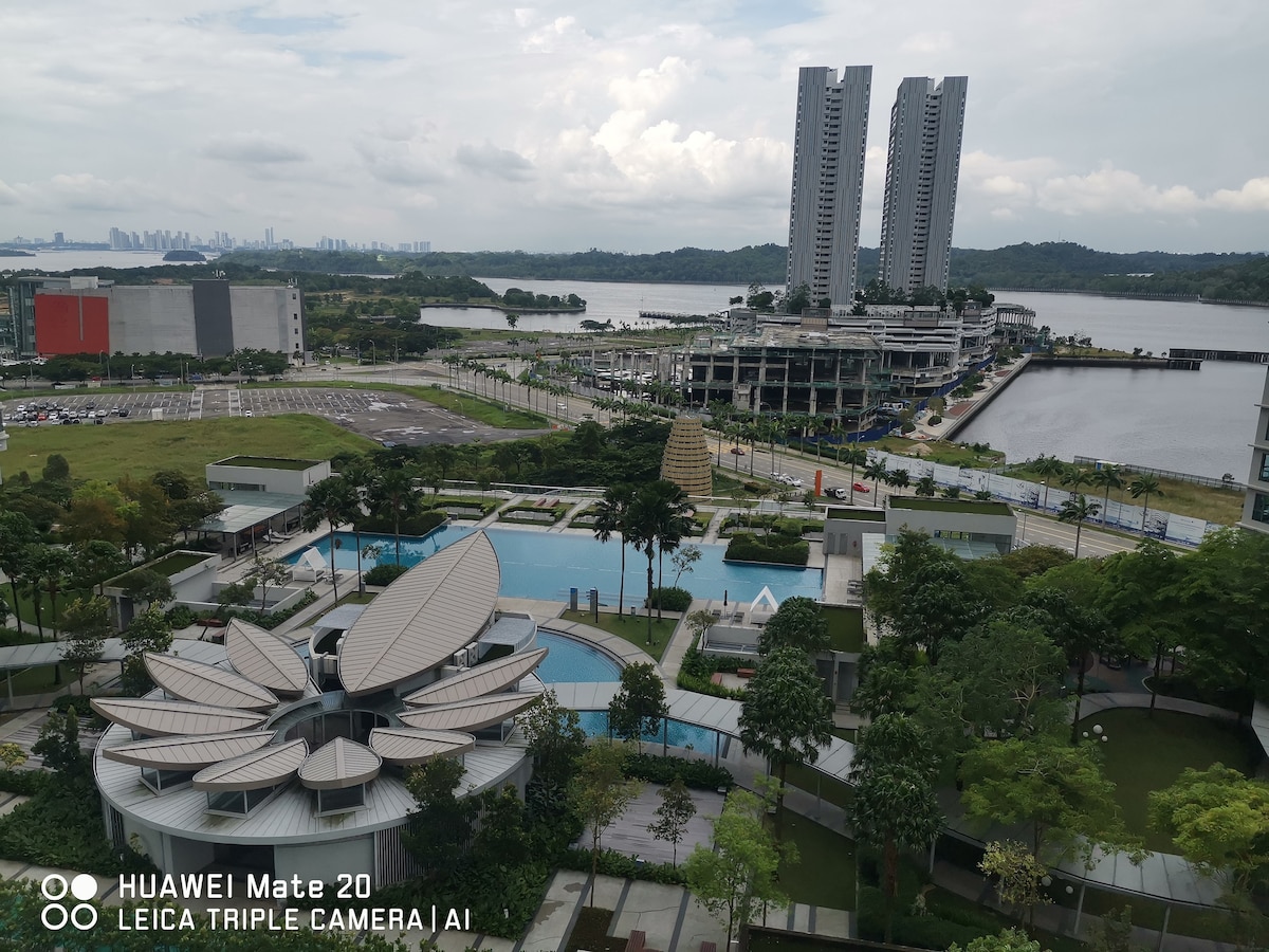 An expansive view of the surrounding area showcases lush greenery and a serene waterfront. A modern pool area with unique architecture is visible, along with residential buildings in the background. The distant skyline hints at a vibrant city atmosphere.