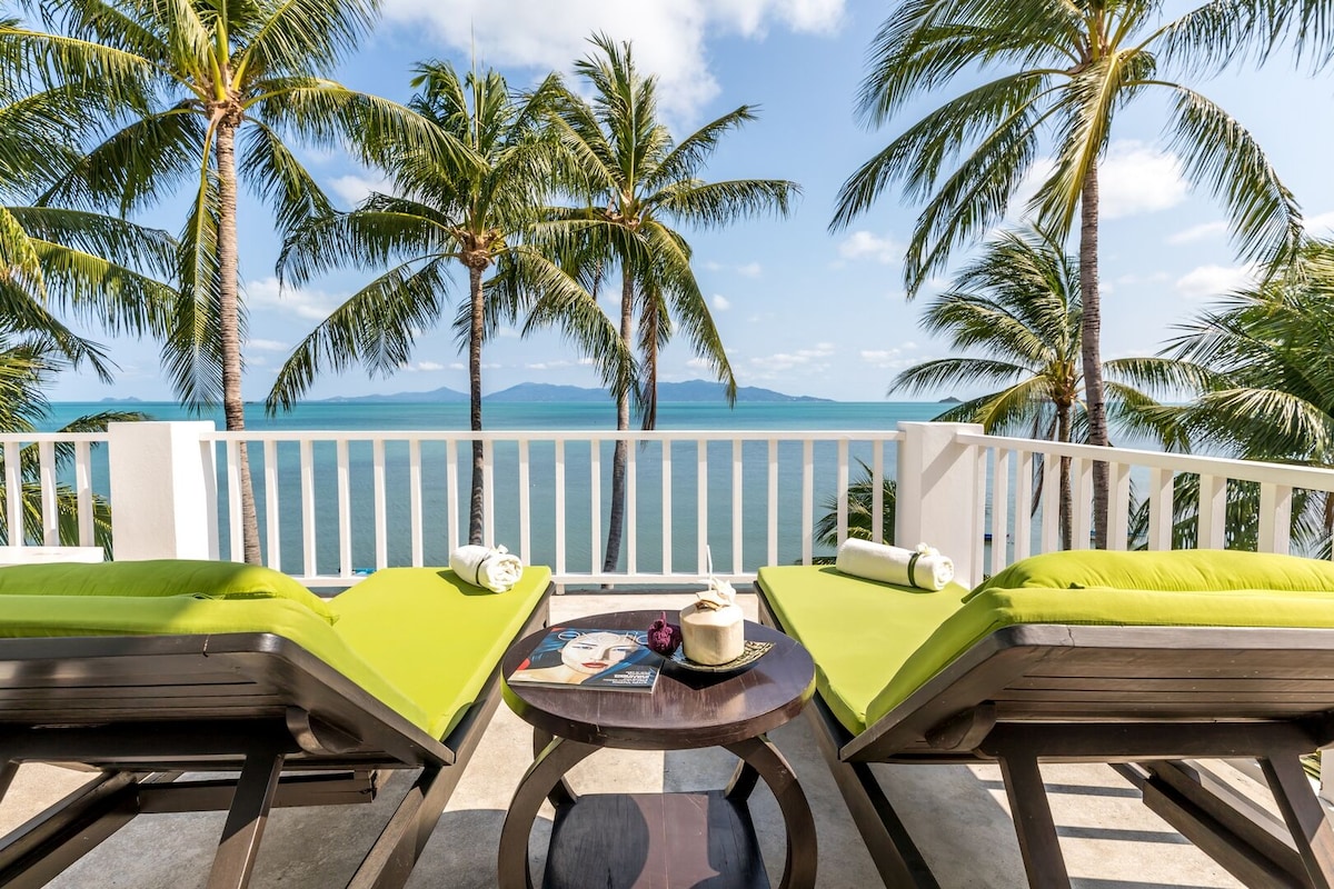 Two sun loungers with bright green cushions are positioned on a balcony, facing views of the vibrant turquoise waters and palm trees swaying in the gentle breeze. A small table holds a magazine and a decorative item, set against a backdrop of distant islands and a clear blue sky.