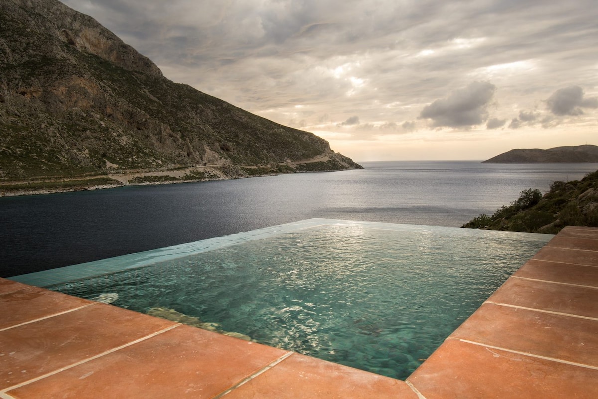 An infinity pool overlooks the serene waters of the bay, framed by rugged hills and cliffs. The tranquil scene is accentuated by a cloudy sky, creating a calm and reflective atmosphere that invites relaxation.