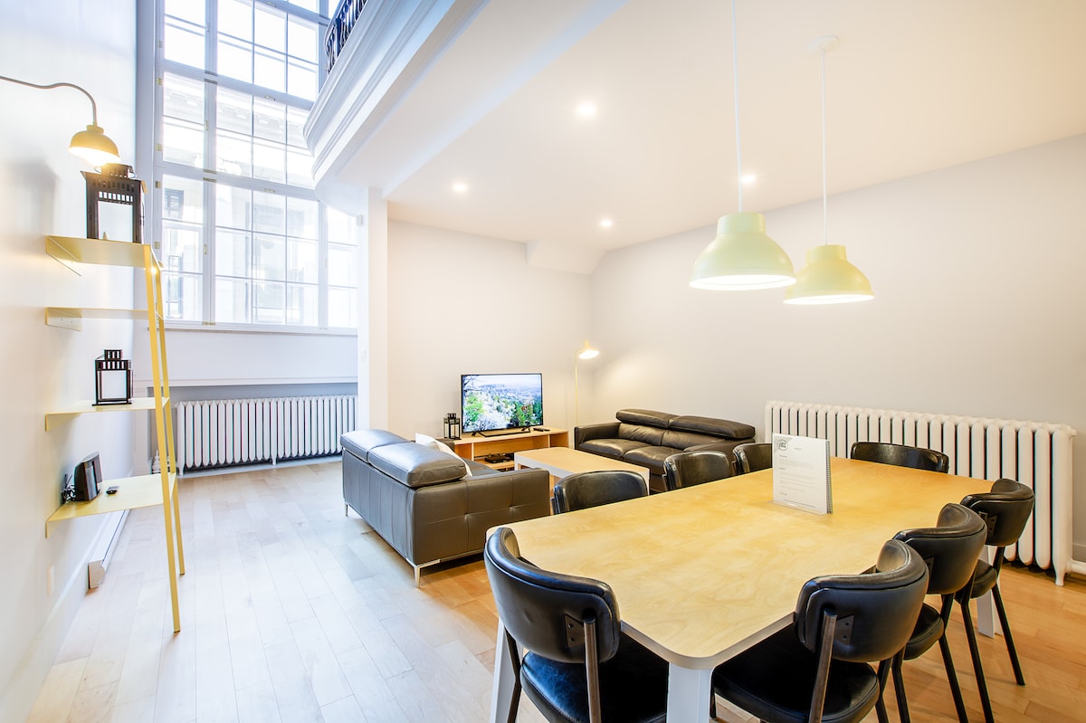 A spacious living area features a large flat-screen TV, two dark leather sofas, and a wooden dining table surrounded by six black chairs. Natural light floods in through tall windows, complemented by modern lighting fixtures hanging above the table.