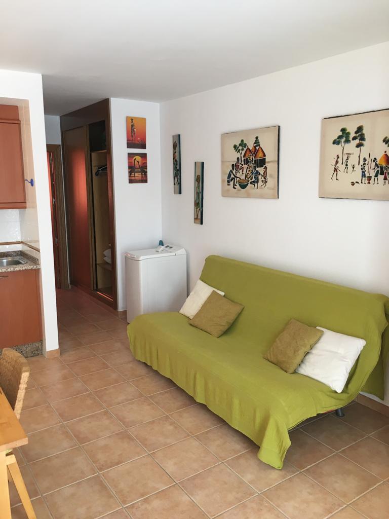 A bright living area features a green sofa bed with decorative pillows. Wall art adds interest to the space, while a table with wicker chairs is positioned nearby. A corridor leads to additional rooms, and natural light illuminates the tiled floor.