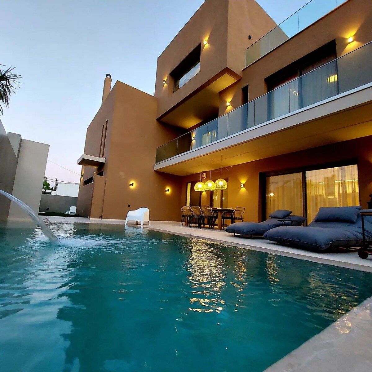 A private heated pool is featured in the foreground, accompanied by loungers and outdoor seating arranged beneath pendant lighting. The villa's modern architecture is showcased in the background, with large windows allowing natural light into the living space.