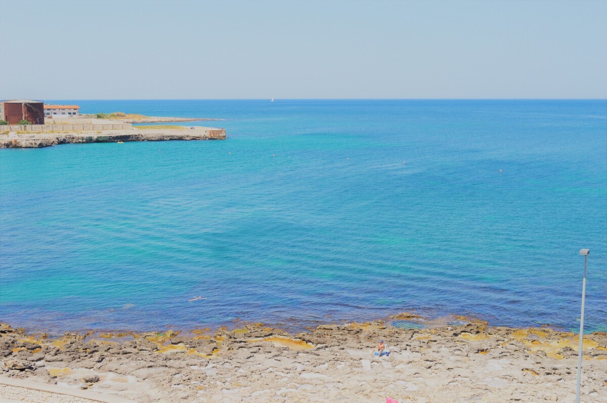 The image captures a clear view of the serene coastline, where turquoise waters gently meet rocky shorelines. The horizon is visible, with a few boats dotting the distant sea, adding to the tranquil seaside atmosphere.