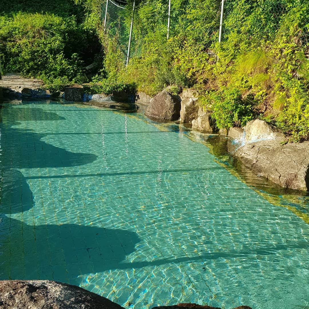 A natural pool is surrounded by rocks and lush greenery. Clear water reflects sunlight, creating shimmering patterns on the surface. Vegetation lines the edges, enhancing the serene atmosphere of the space.
