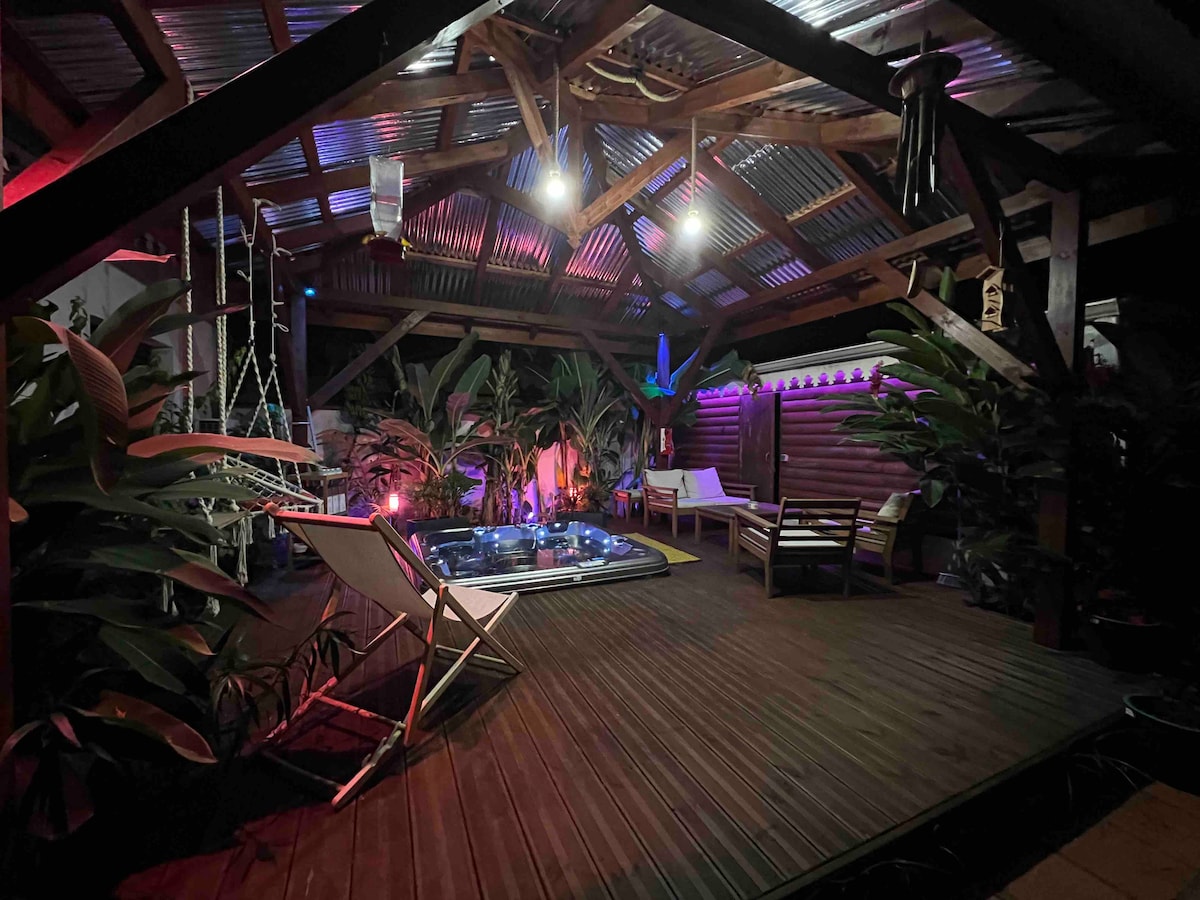 An inviting outdoor area features a private jacuzzi surrounded by lush tropical plants. Comfortable lounge chairs are positioned nearby, and the space is enhanced by soft lighting that highlights the wooden structure and ceiling beams.