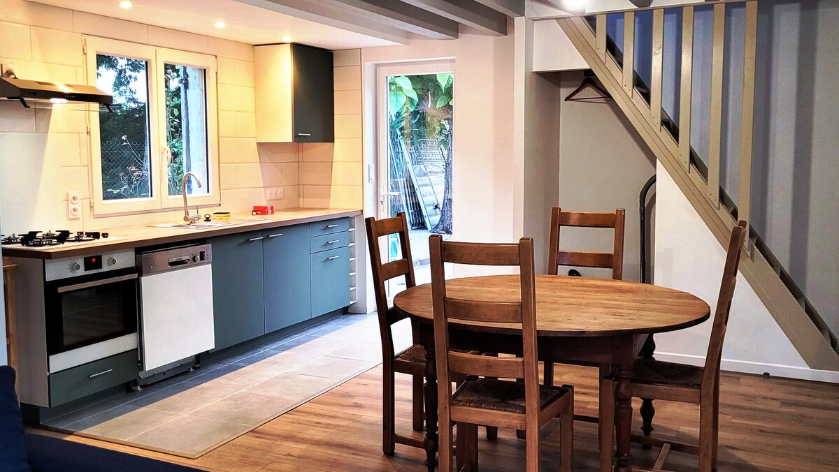 An open living area is shown, featuring a circular wooden dining table with several wooden chairs. A well-equipped kitchen is visible, showcasing modern appliances and cabinetry, with natural light flooding in from large windows. Stairs lead to a private upper level.