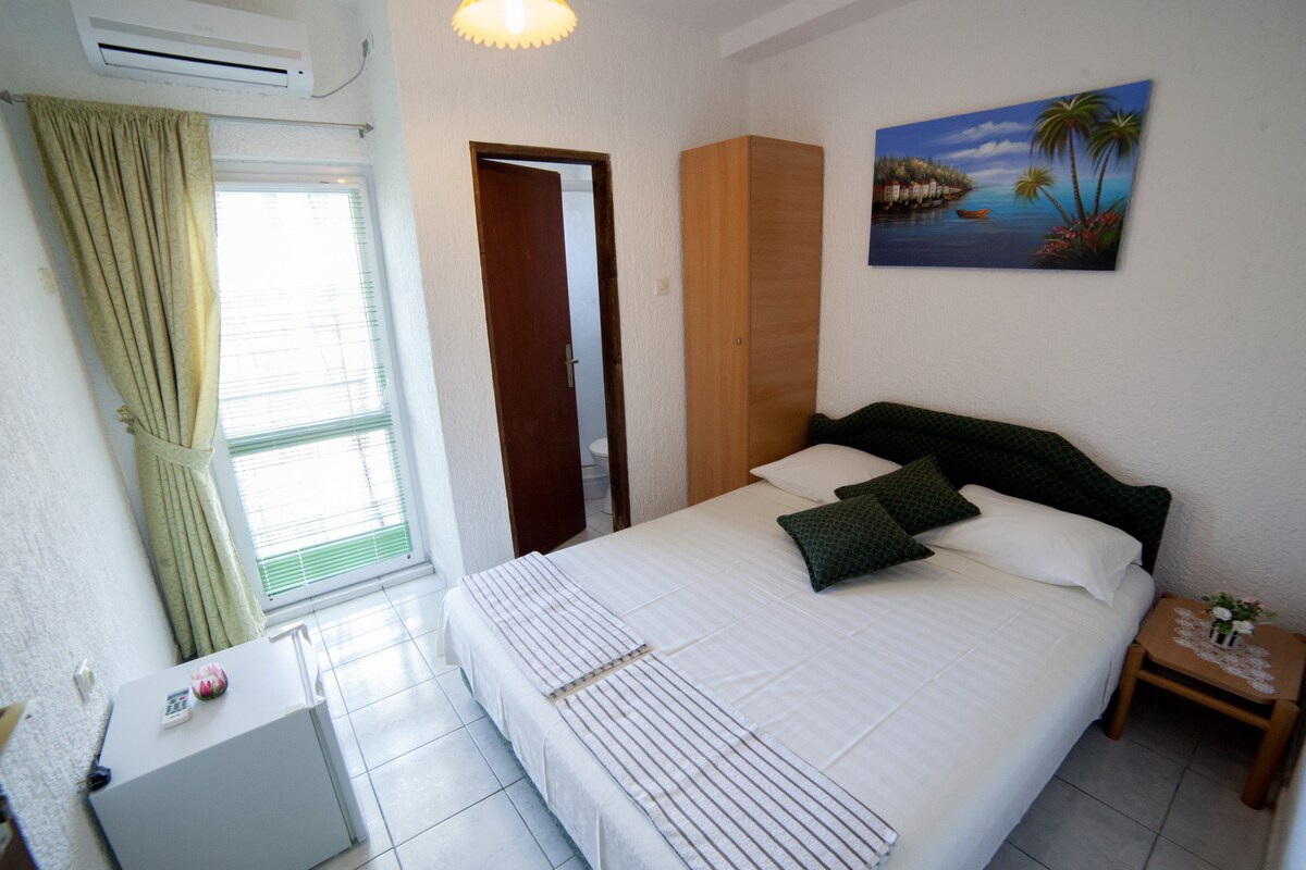 A bright sleeping area features a spacious bed with soft linens and decorative pillows. A small table holds a potted plant, while a window allows natural light to fill the room. Light-colored walls and a painting of a tropical scene enhance the calming decor.